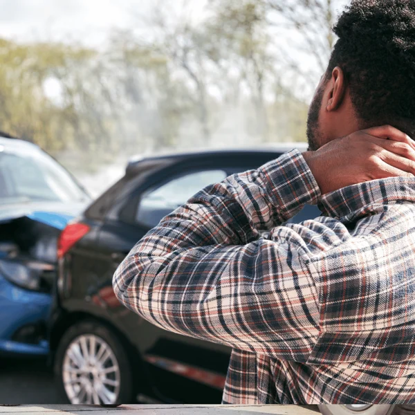 A man holds his neck while looking at two cars involved in a rear-end collision on a road during daytime.