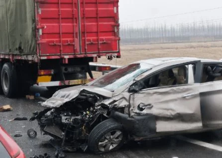 A silver car with severe front and side damage is stopped near the rear of a red truck on a wet road; debris is scattered around.