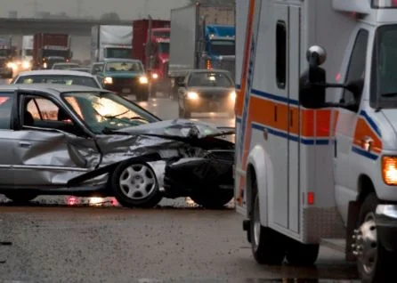 A damaged car and an ambulance are stopped on a wet, busy highway during a traffic accident. Other vehicles are visible in the background.