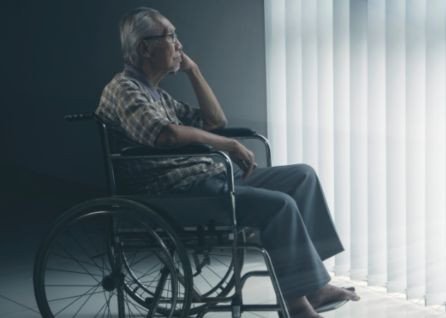 An elderly person in a wheelchair sits indoors, looking out a window with vertical blinds, appearing thoughtful.