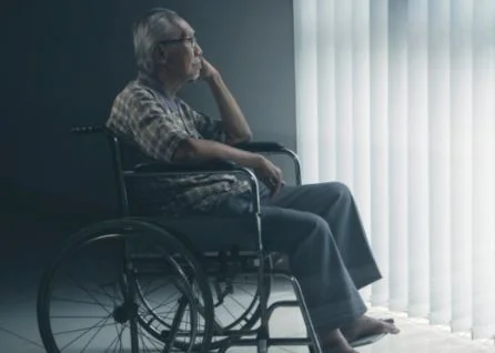 An elderly person in a wheelchair sits indoors, looking out a window with vertical blinds, appearing thoughtful.