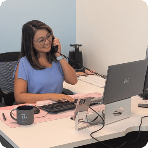 Woman wearing glasses and a blue shirt sits at a desk, speaking on a corded phone while typing on a keyboard, with a mug and smartphone nearby.