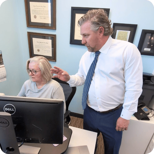A man in a white shirt and tie stands beside a seated woman at a desk, looking at her computer screen in an office with framed certificates on the wall.