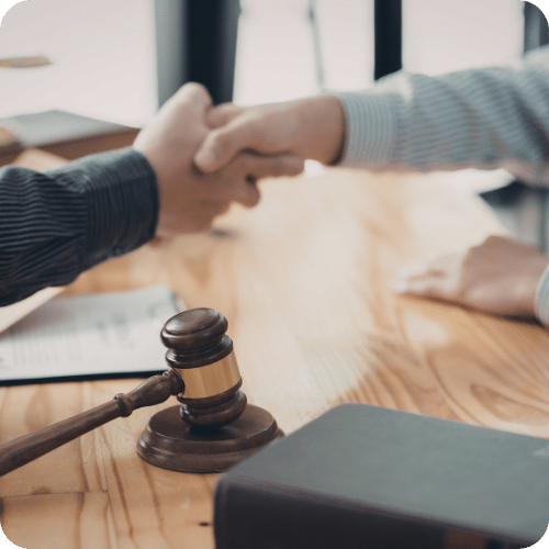Two people shake hands across a wooden table with a gavel and documents, suggesting a legal agreement or settlement.