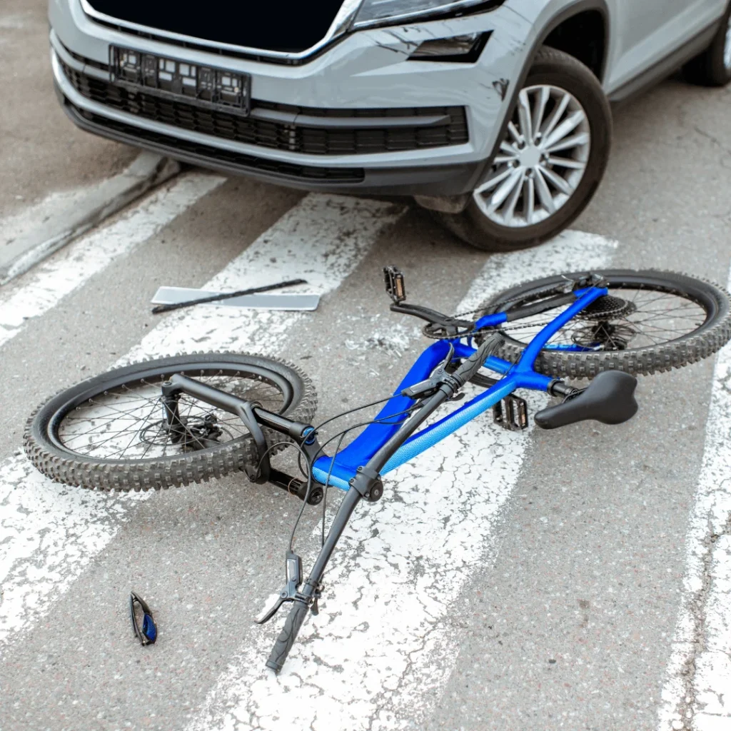 A blue bicycle lies on a crosswalk in front of a gray car with a bent wheel, suggesting a recent collision; sunglasses are on the ground nearby.