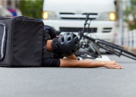 A cyclist wearing a helmet and backpack lies on the road next to a fallen bicycle, with a white van stopped behind them.