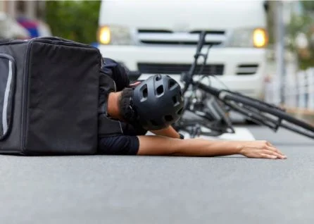 A cyclist wearing a helmet and backpack lies on the road next to a fallen bicycle, with a white van stopped behind them.