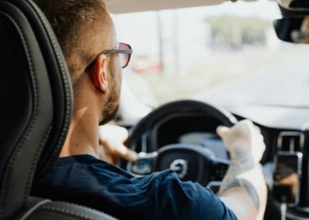 A man with short hair and glasses drives a car, viewed from the back seat during the daytime.