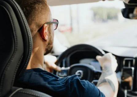 A man with short hair and glasses drives a car, viewed from the back seat during the daytime.
