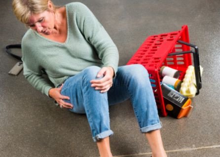 A woman sits on the floor holding her knee beside a fallen red shopping basket with groceries spilled out.