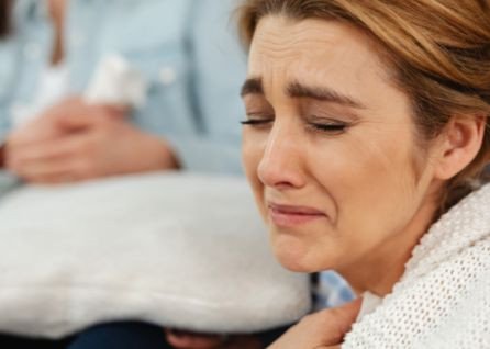 Close-up of a woman crying with a distressed facial expression, holding her hand to her chest. Another person is blurred in the background holding a pillow.