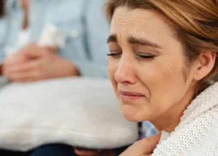 Close-up of a woman crying with a distressed facial expression, holding her hand to her chest. Another person is blurred in the background holding a pillow.